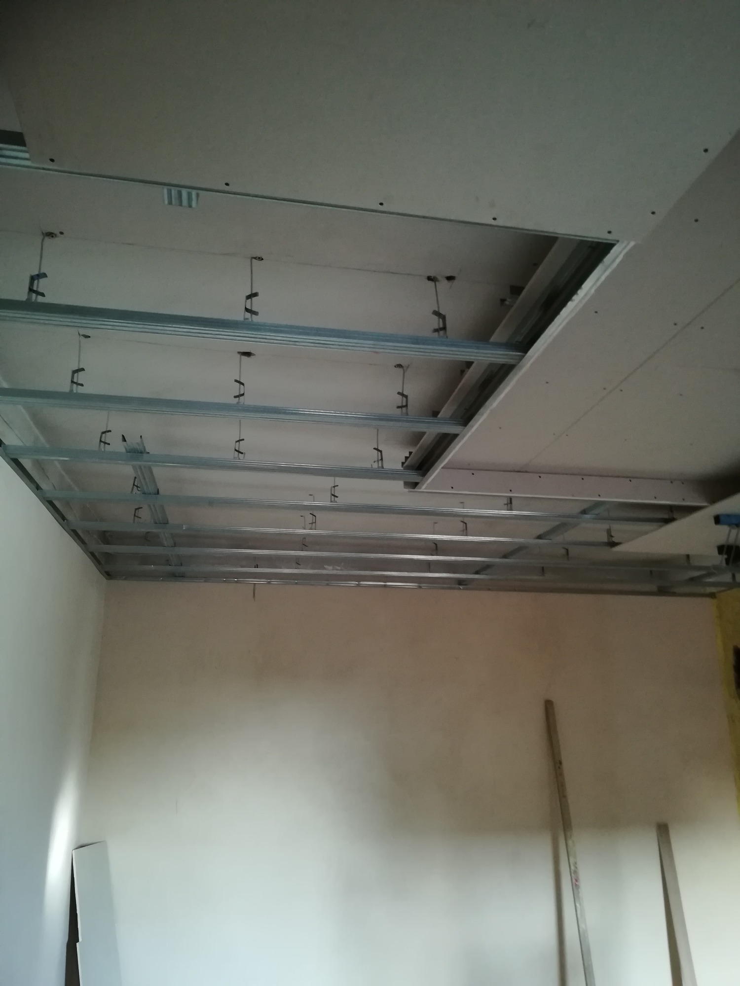 Montaż sufitu podwieszanego z płyt kartonowo-gipsowych, widoczna metalowa konstrukcja nośna i częściowo zamontowane płyty, ściany w stanie surowym.