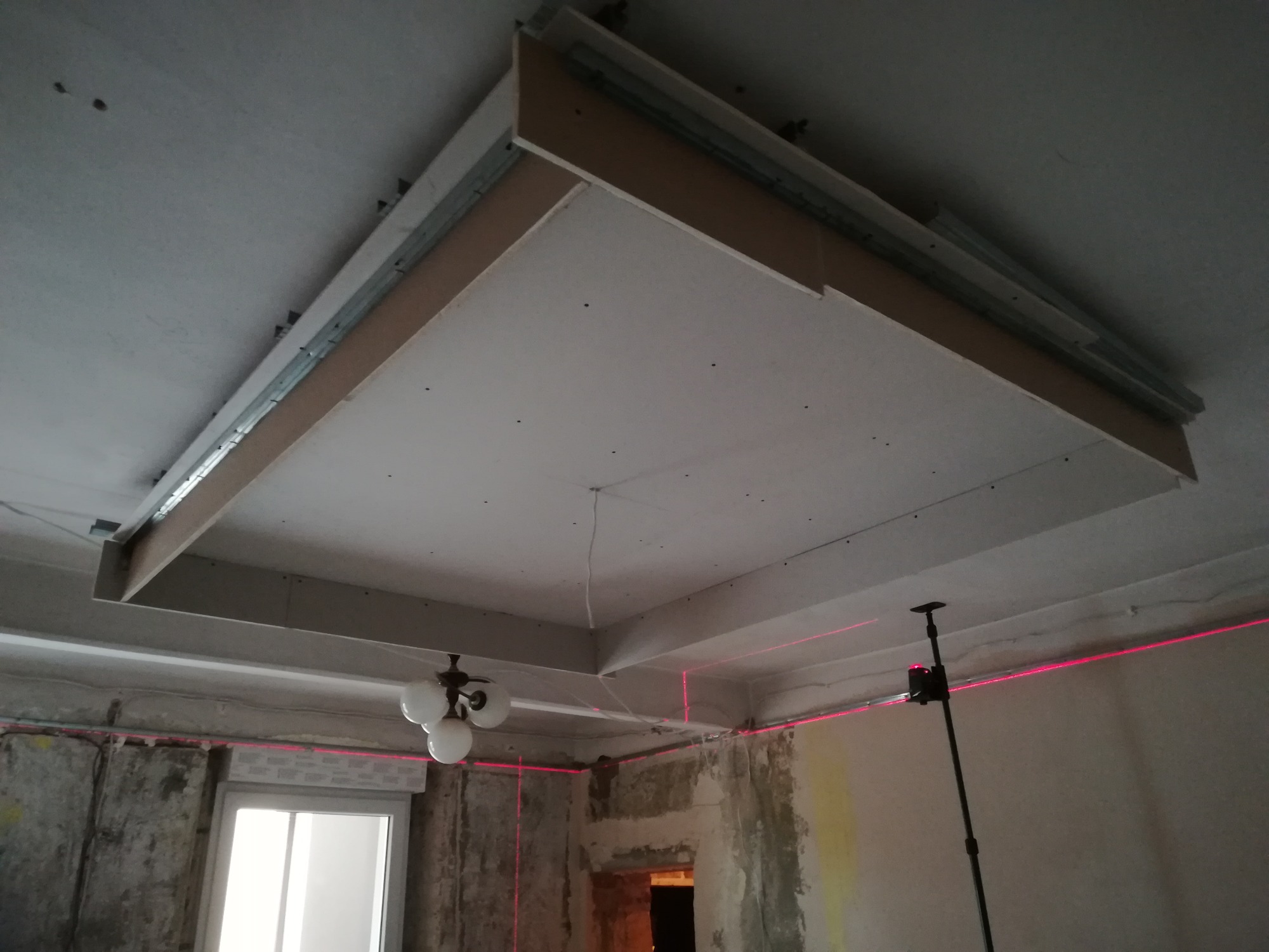 Montaż podwieszanego sufitu z płyt kartonowo-gipsowych, widoczne profile metalowe, płyty oraz laserowy niwelator wyznaczający poziom na ścianach w pomieszczeniu w trakcie prac.