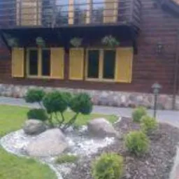 Ogród przydomowy z ozdobnymi krzewami i kamieniami, na tle drewnianego domu z balkonem i żółtymi okiennicami. Aranżacja rabaty z obrzeżem.