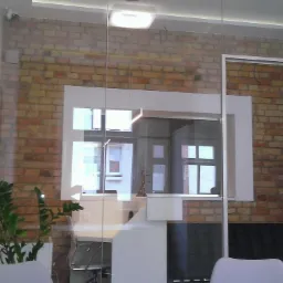 Nowoczesne biuro z ceglaną ścianą, przeszklonymi drzwiami i widoczną rośliną w doniczce. Minimalistyczny design, białe meble, naturalne światło.