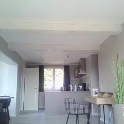 Jasne, drewniane belki stropowe w minimalistycznym wnętrzu z szarą kuchnią w tle, widok z niskiego kąta.