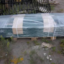 Paleta z zielonymi panelami ogrodzeniowymi owiniętymi folią, zabezpieczonymi taśmą, z fragmentem dekoracyjnego elementu metalowego ogrodzenia w tle, na ziemi z roślinnością.