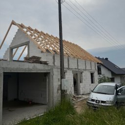 ZAKŁAD OGÓLNOBUDOWLANY ANDRZEJ KOSZAŁKA - Surowy dom z bloczków z drewnianą konstrukcją dachu w trakcie budowy, widok z zewnątrz, samochód obok. Widoczne elementy instalacji elektrycznej na ścianie.