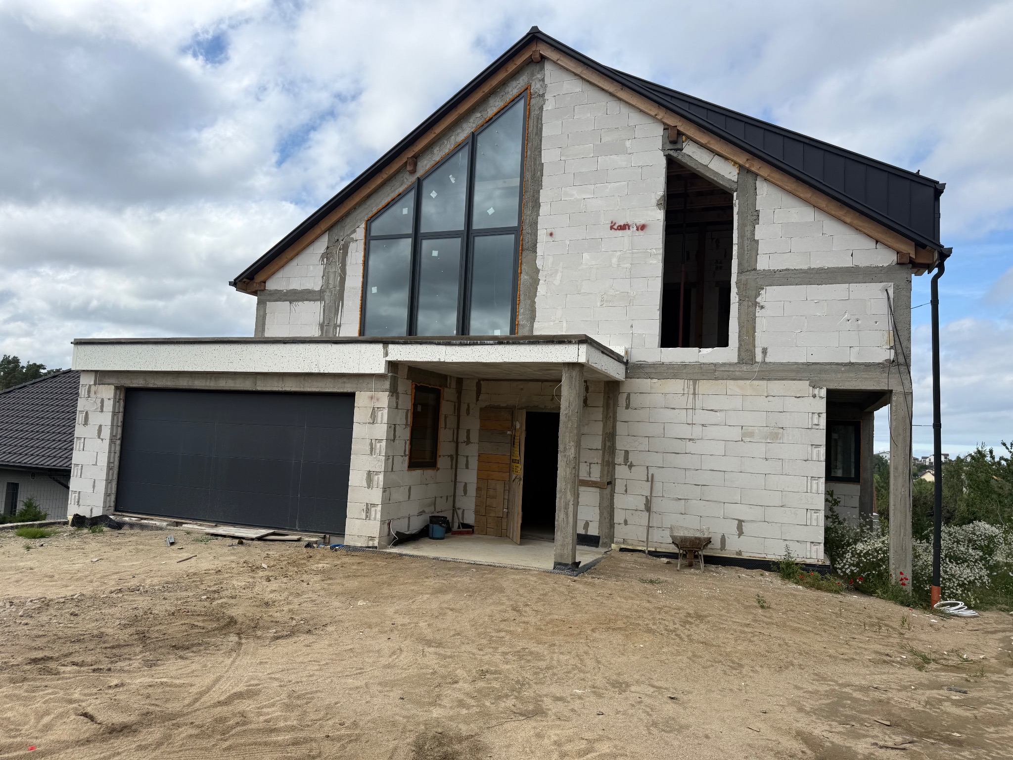 Surowy dom z białego betonu komórkowego w trakcie budowy, z zamontowanymi czarnymi oknami i garażem. Widoczne zarysowane kontury okien i drzwi.