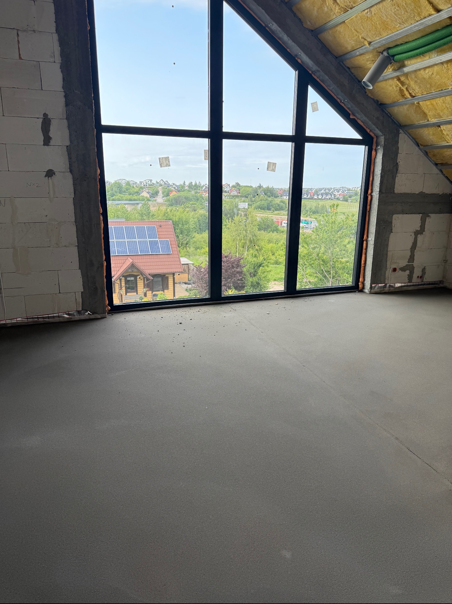 Wnętrze poddasza z dużym oknem, widok na dom z panelami słonecznymi. Ściany z białego betonu komórkowego, ocieplenie poddasza żółtą wełną, wylewka podłogowa.