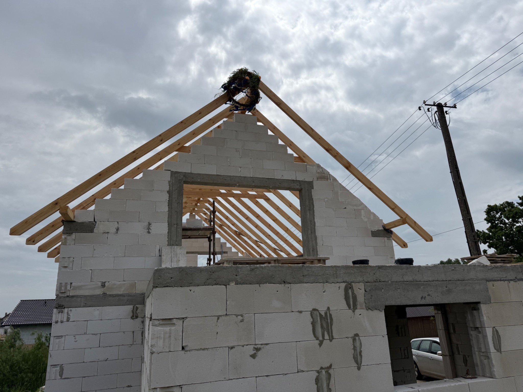 Niedokończony dom z białych pustaków z wieńcem na dachu w słoneczny dzień, widok na drewnianą konstrukcję dachu i okna.