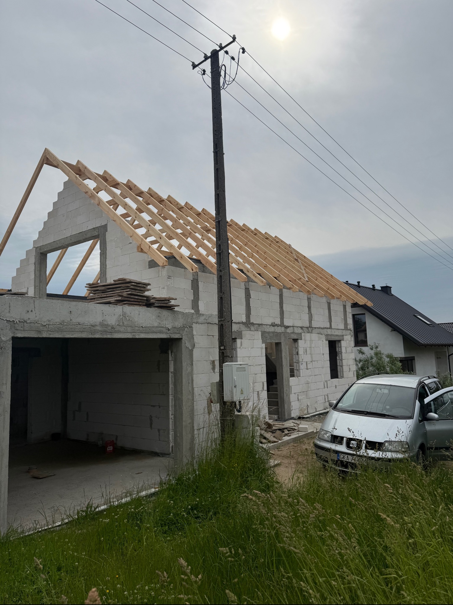 Surowy dom z bloczków z drewnianą konstrukcją dachu w trakcie budowy, widok z zewnątrz, samochód obok. Widoczne elementy instalacji elektrycznej na ścianie.