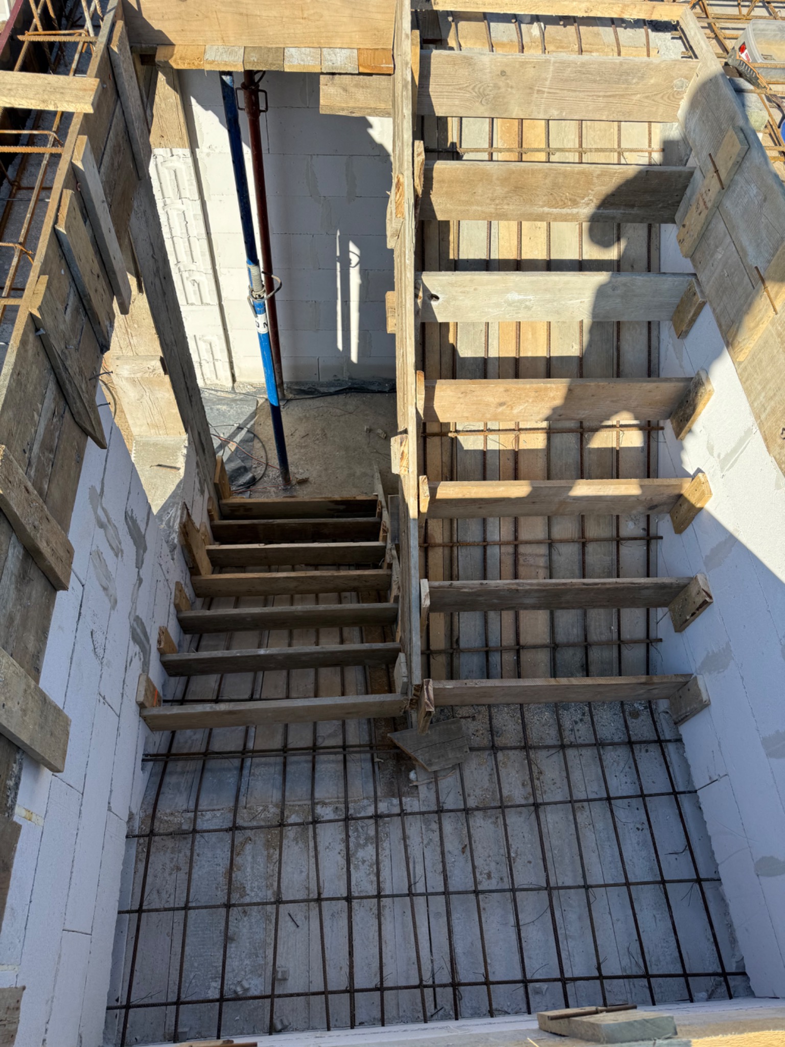 Szkielet betonowych schodów z drewnianym szalunkiem i zbrojeniem, wewnątrz ścian z betonu komórkowego. Widok z góry na budowę.