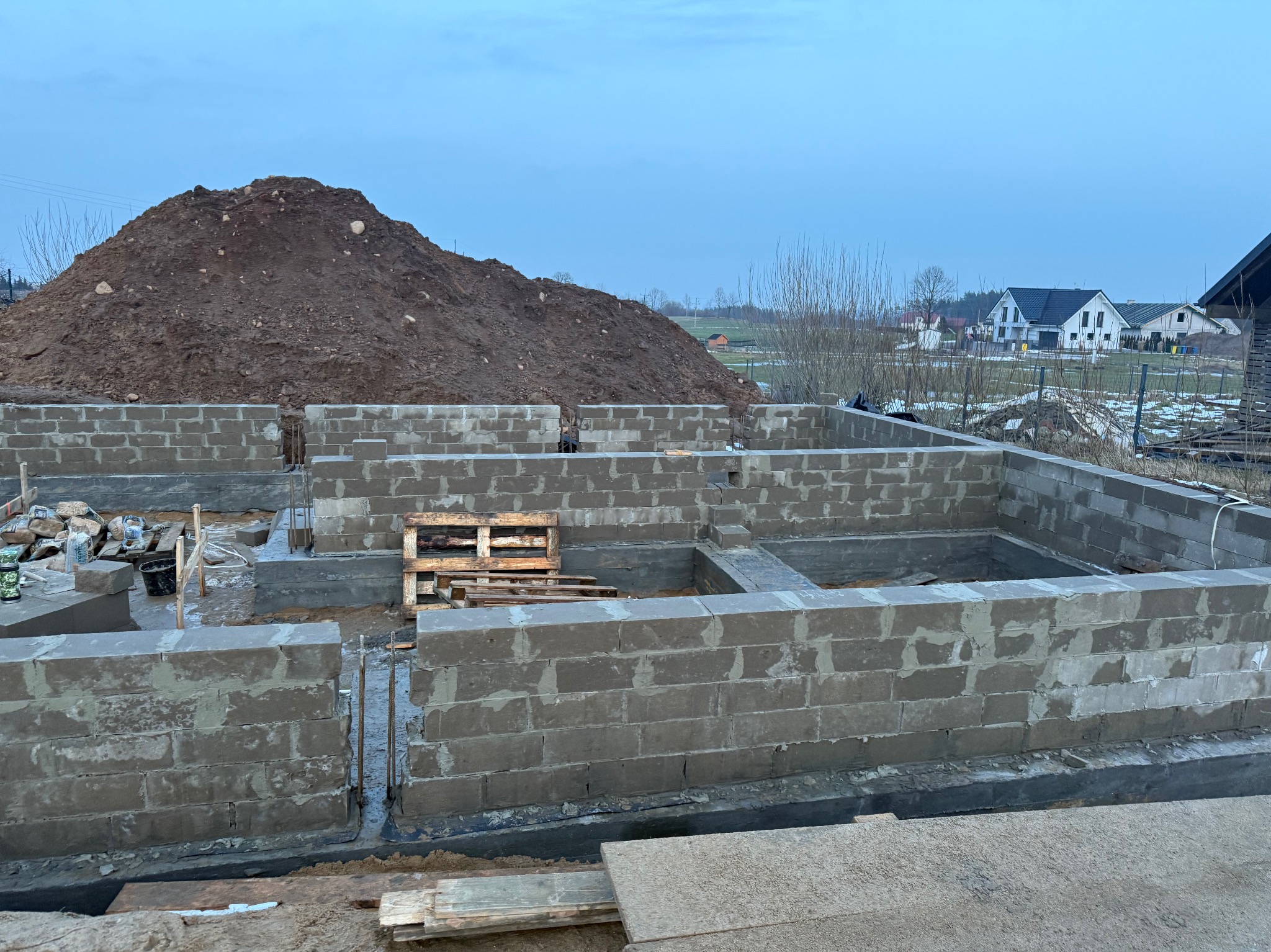 Budowa fundamentów domu z bloczków betonowych, widoczny wykop i sterta ziemi. Na drugim planie zabudowa wiejska i pochmurne niebo.