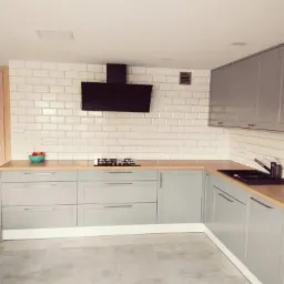 Jasna kuchnia w stylu skandynawskim z szarymi szafkami, drewnianym blatem, białymi płytkami na ścianie i czarnym okapem.