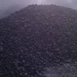 Kopalnia: Duża pryzma antracytu w metalowej skrzyni, widoczny fragment ceglanej ściany w tle. Niejednolita struktura węgla.