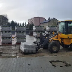 Żółta ładowarka Volvo transportuje paletę kostki brukowej na placu budowy, w tle stosy kostki i budynki mieszkalne w pochmurny dzień.