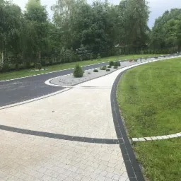 Kremowa kostka brukowa ułożona w wzór jodełki, obramowana ciemnoszarą kostką, tworząca ścieżkę przez trawnik i łącząca się z czarną nawierzchnią z kostki, otoczoną rabatami z kamyków i niskimi...