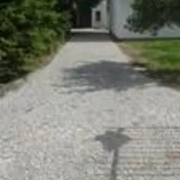 Jasnoszara ścieżka z drobnej kostki prowadząca do budynku, oświetlona słońcem, z widocznym cieniem osoby robiącej zdjęcie. Otoczenie: trawa i drzewa.