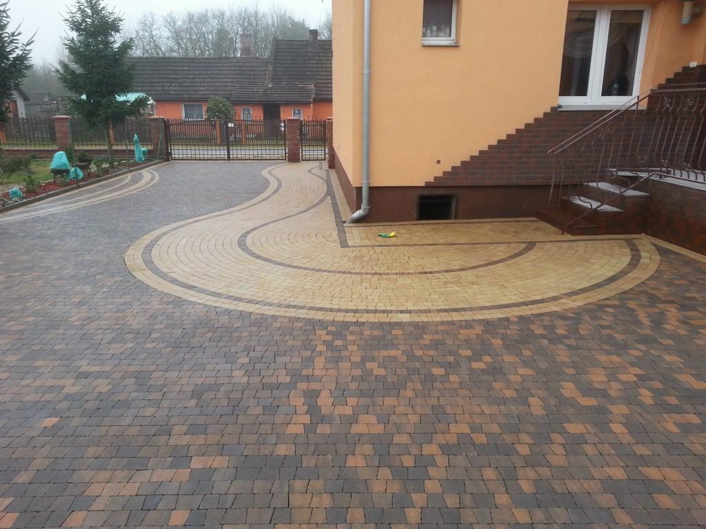 Ułożona, dwukolorowa kostka brukowa tworząca wzór na podjeździe. Elementy dekoracyjne z kostki w kształcie łuku przy schodach budynku. Widok na posesję.