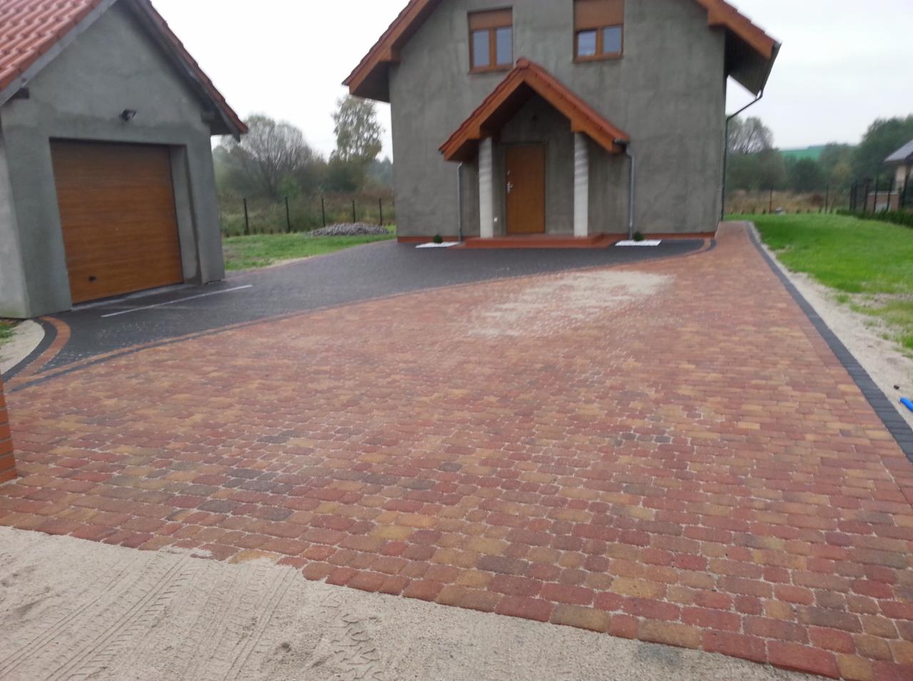 Ułożona kostka brukowa w odcieniach czerwieni i brązu tworzy wzorzysty podjazd do domu z garażem. Fragment podsypki piaskowej widoczny w dolnym rogu.
