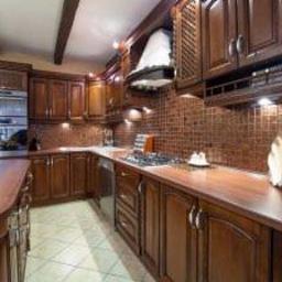 Sklep Meblowy - Klasyczna kuchnia z ciemnego drewna, brązowe płytki na ścianie, beżowa podłoga w kratkę, oświetlenie punktowe pod szafkami, drewniane belki na suficie.