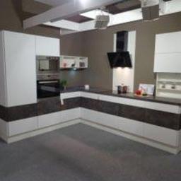 Sklep Meblowy - Nowoczesna kuchnia w kształcie litery L z białymi szafkami górnymi i dolnymi, ciemnym blatem oraz brązowym pasem dekoracyjnym. Widoczny piekarnik, mikrofalówka i okap. Oświetlenie punktowe...