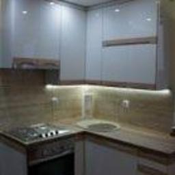 Sklep Meblowy - Nowoczesna, narożna zabudowa kuchenna z białymi, lakierowanymi frontami i drewnopodobnym blatem, oświetlenie LED podszafkowe i punktowe, płyta gazowa zintegrowana z blatem.