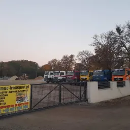 Plac firmy transportowej z zaparkowanymi ciężarówkami MAN, stertami piasku i żwiru w tle oraz banerem reklamowym na ogrodzeniu.
