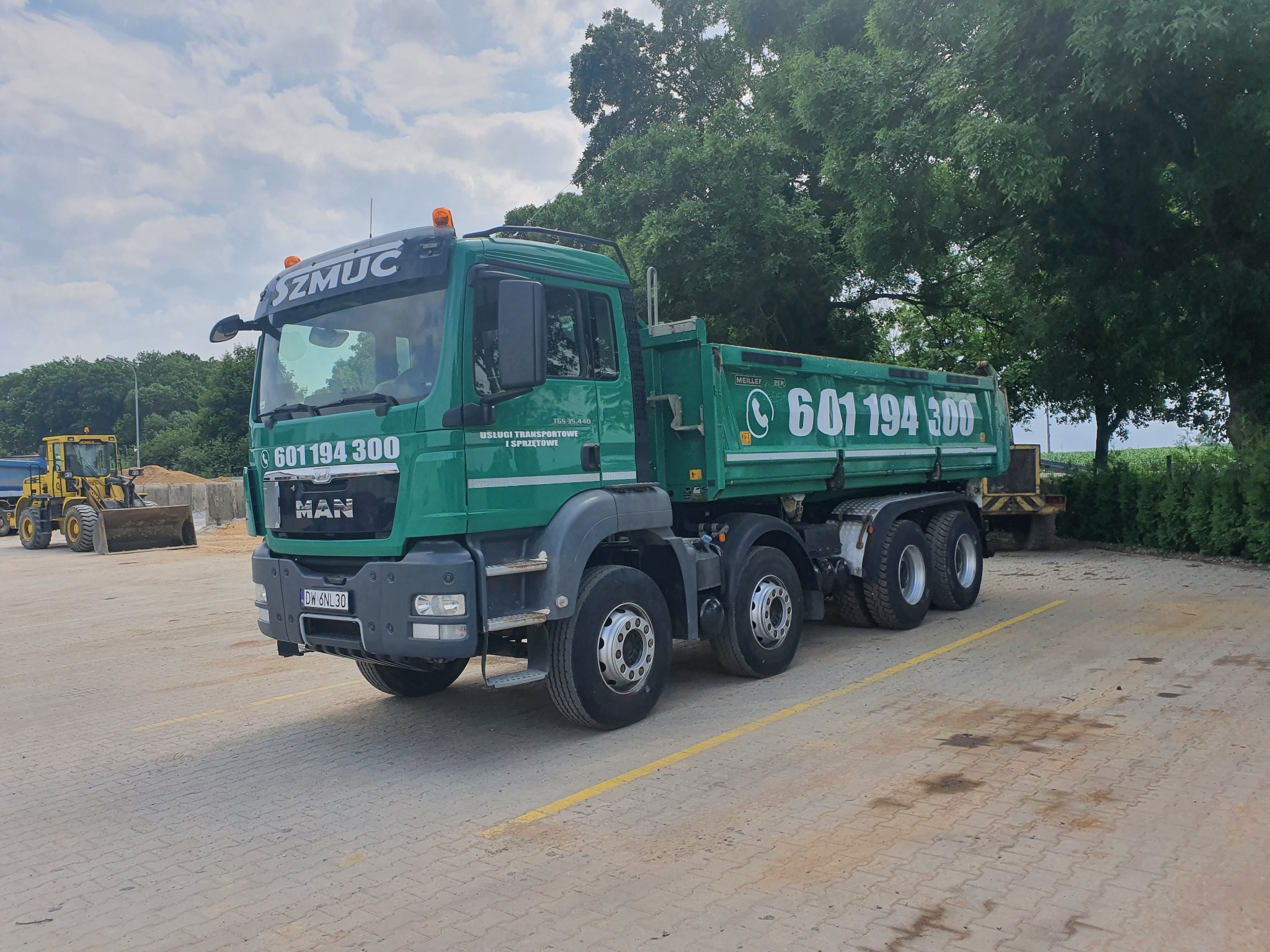 Zielona ciężarówka MAN z numerem telefonu firmy transportowej na naczepie, zaparkowana na placu z kostki brukowej, w tle widoczna ładowarka.