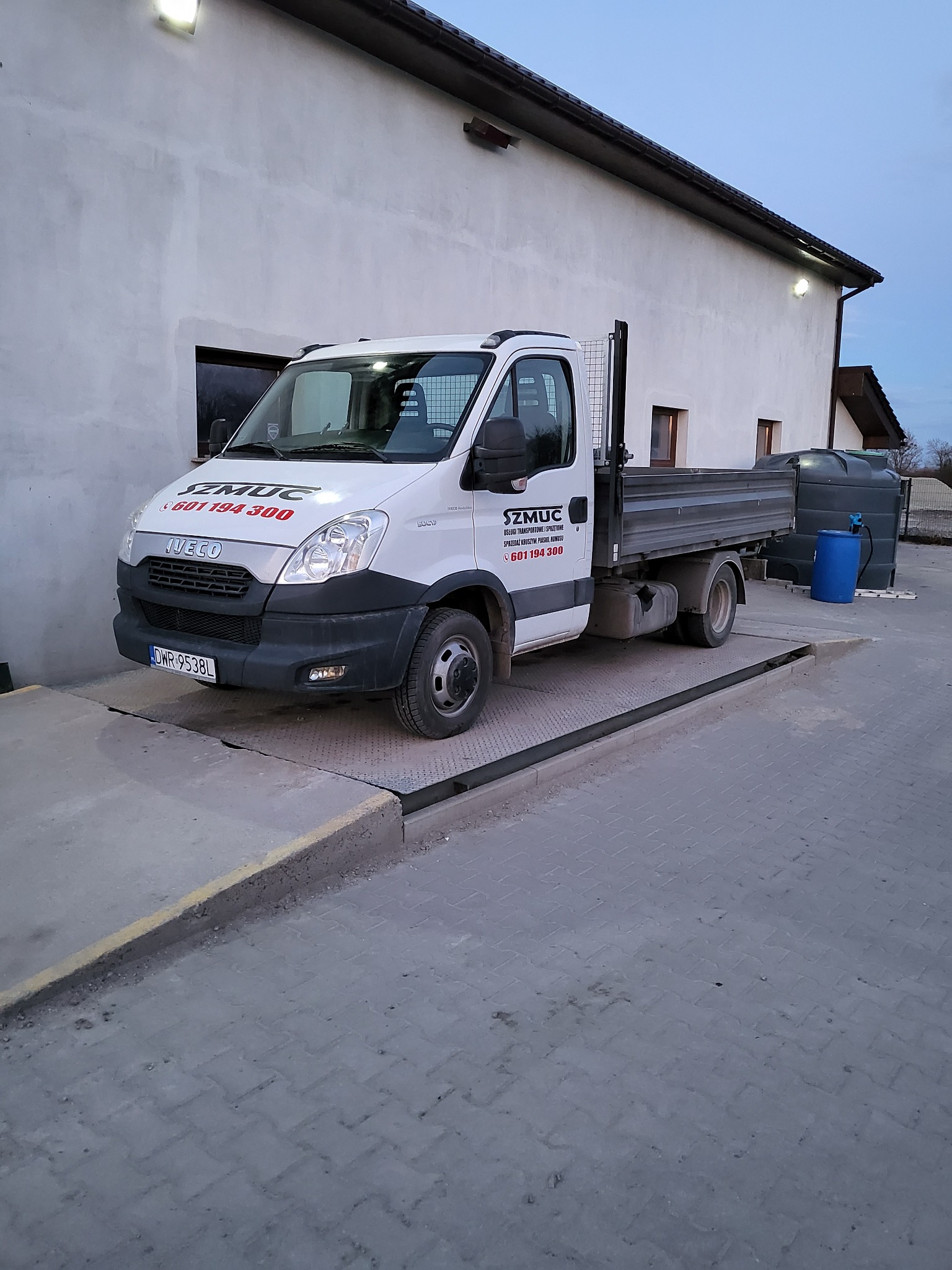 Biały samochód ciężarowy marki Iveco z otwartą skrzynią ładunkową, zaparkowany na betonowej platformie przed budynkiem z białymi ścianami. Na pojeździe widoczne logo firmy 'SZMUC' oraz numer...