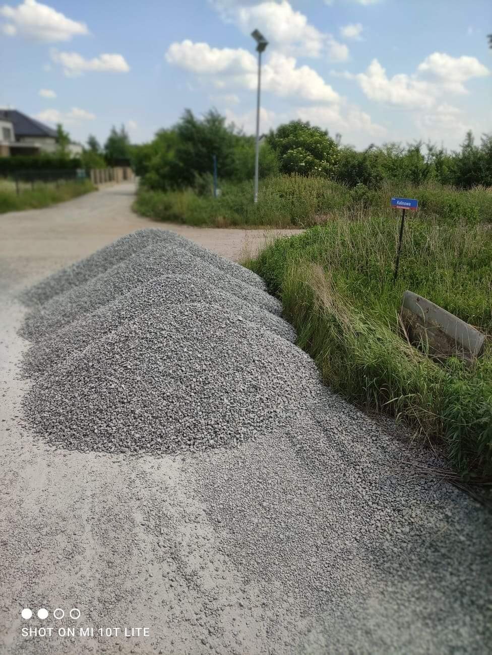 Kupa szarego kruszywa na piaszczystej drodze wiejskiej, obok tabliczka z nazwą ulicy i wysoka trawa, w tle domy i drzewa.