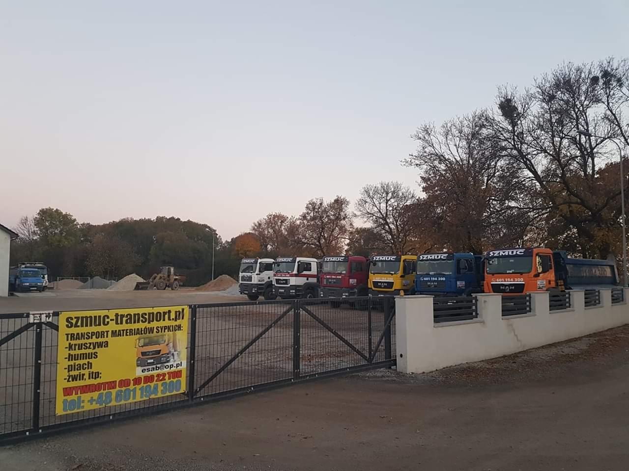 Plac firmy transportowej z zaparkowanymi ciężarówkami MAN, stertami piasku i żwiru w tle oraz banerem reklamowym na ogrodzeniu.