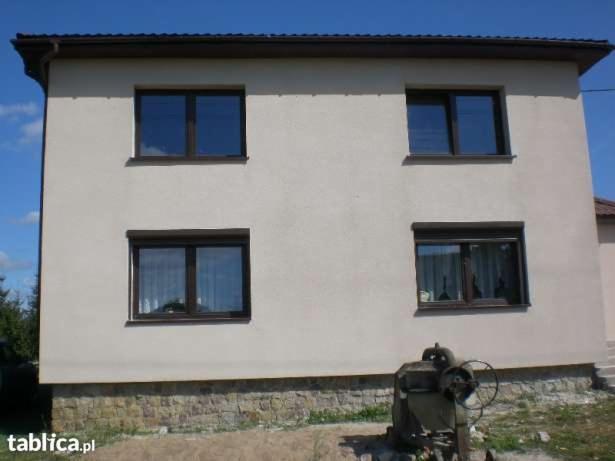 Dwukondygnacyjny dom z jasną elewacją, brązowymi ramami okiennymi i kamienną podmurówką, z widoczną starą pompą wodną na pierwszym planie.