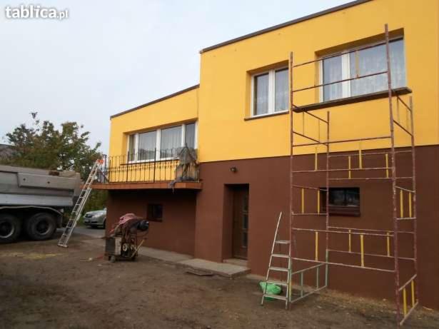 Dwukondygnacyjny dom z żółtą elewacją górnej części i brązową dolnej, z rusztowaniem przy ścianie i betoniarką na podwórku.