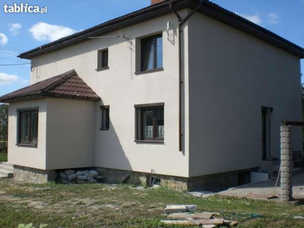 Dwukondygnacyjny dom z jasną elewacją, brązowymi ramami okiennymi i dachem z brązowej dachówki, widoczny z zewnątrz w słoneczny dzień.