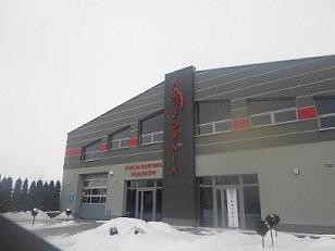 Budynek z szarymi panelami i czerwonymi akcentami, z napisem 'IZOLOWNIA' i logiem w kształcie płomienia na pionowym panelu. Przed budynkiem leży śnieg.