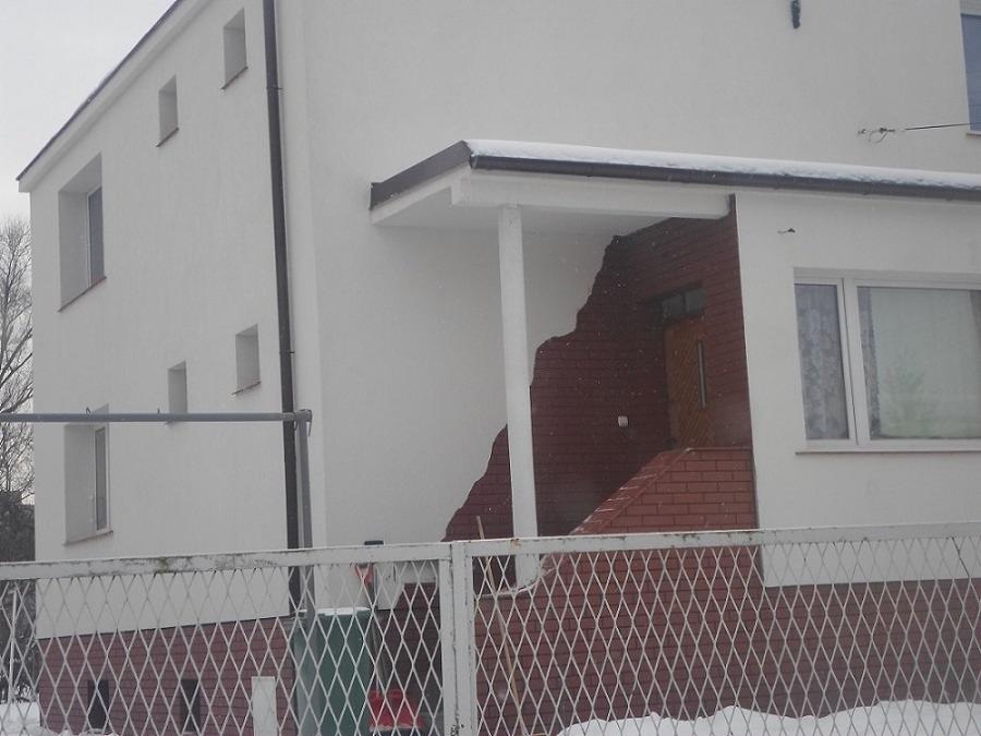 Dwupiętrowy dom z białymi ścianami i ceglanym wejściem, widziany zza metalowego ogrodzenia w pochmurny dzień.