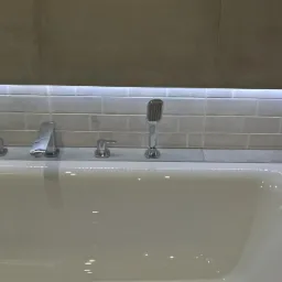 Biała wanna z chromowaną armaturą i słuchawką prysznicową, podświetlona listwą LED, wykończona szarymi płytkami typu cegiełka.