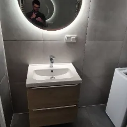 Nowoczesna łazienka z szarymi płytkami, okrągłe lustro z podświetleniem LED, biała umywalka z drewnianą szafką i widoczne odbicie osoby robiącej zdjęcie.