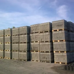 Ułożone na paletach szare bloczki betonowe pod błękitnym niebem. Widoczne ślady opon na piasku. Część bloczków leży luzem na ziemi.