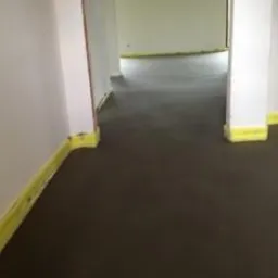 Świeżo wylana, szara posadzka w pomieszczeniu. Ściany i filary zabezpieczone żółtą folią malarską. Widoczny fragment drugiego pomieszczenia w tle.