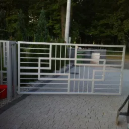 Nowoczesna, metalowa brama wjazdowa o geometrycznym wzorze, pomalowana na srebrno, widoczna z zewnątrz posesji, z czerwoną donicą u podstawy jednego ze słupków.