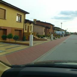 KIAN Kinga Jeziorecka - Ulica osiedlowa z rzędem domów jednorodzinnych, widok z wnętrza samochodu, kostka brukowa w dwóch kolorach, małe drzewka ozdobne przed budynkami.