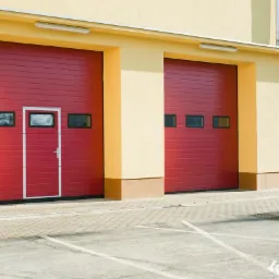 Trzy czerwone segmentowe konstrukcje z oknami i drzwiami w jednej z nich, zamontowane w budynku o żółtej fasadzie, widok z zewnątrz na parkingu.