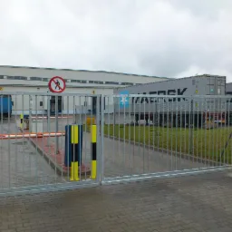 Metalowa brama przesuwna z pionowymi prętami na tle budynku magazynowego i kontenerów transportowych, znak zakazu przejścia pieszych.