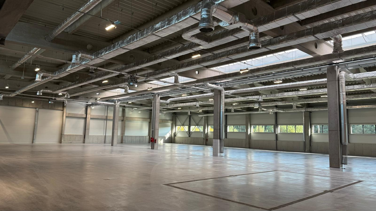 Wnętrze hali produkcyjnej z widocznymi instalacjami wentylacyjnymi pod sufitem i betonową posadzką. Hala oświetlona światłem dziennym wpadającym przez okna.