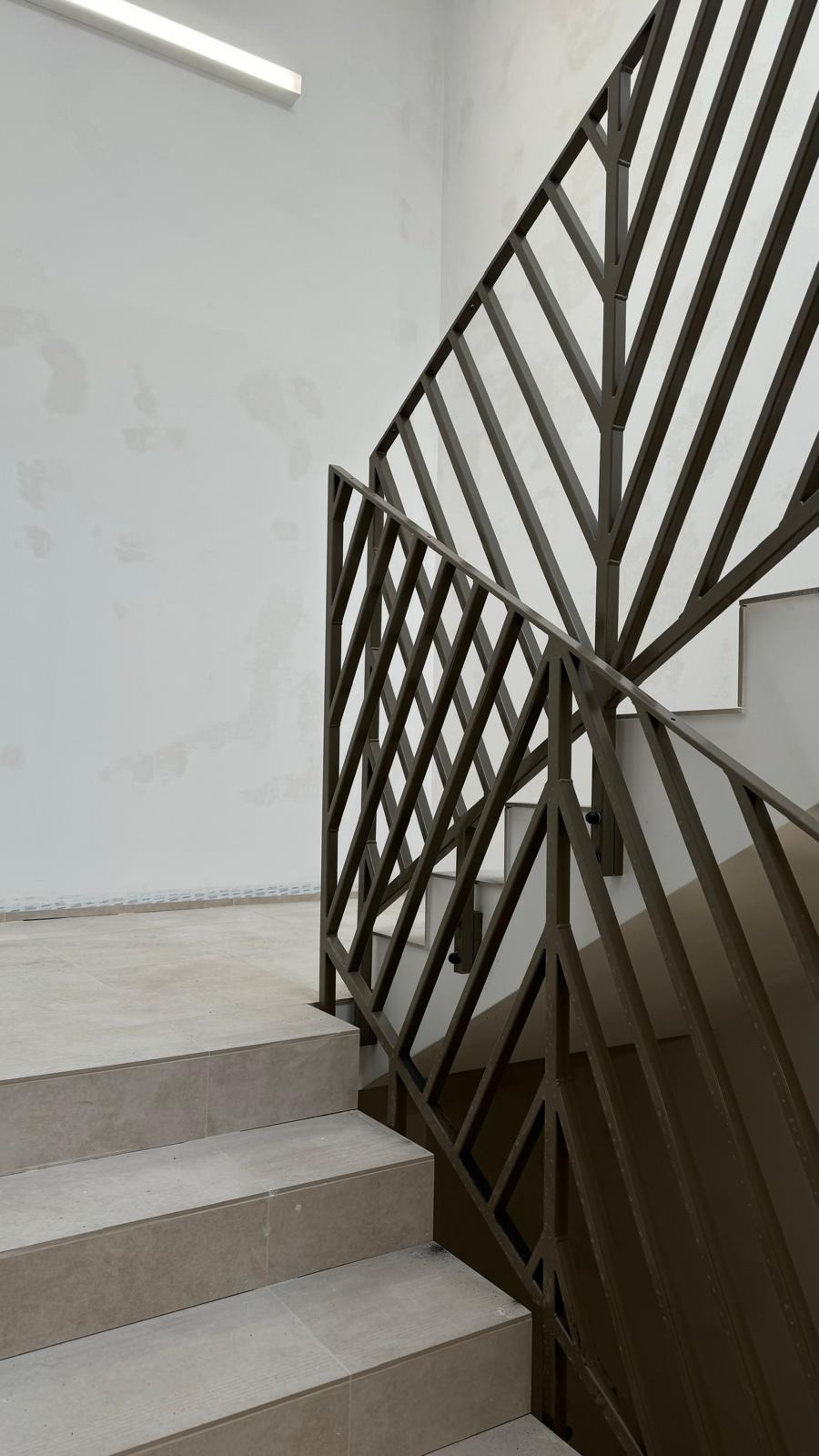 Nowoczesna, metalowa balustrada schodowa z geometrycznym wzorem w brązowym odcieniu, na tle jasnych płytek schodowych i białej ściany.