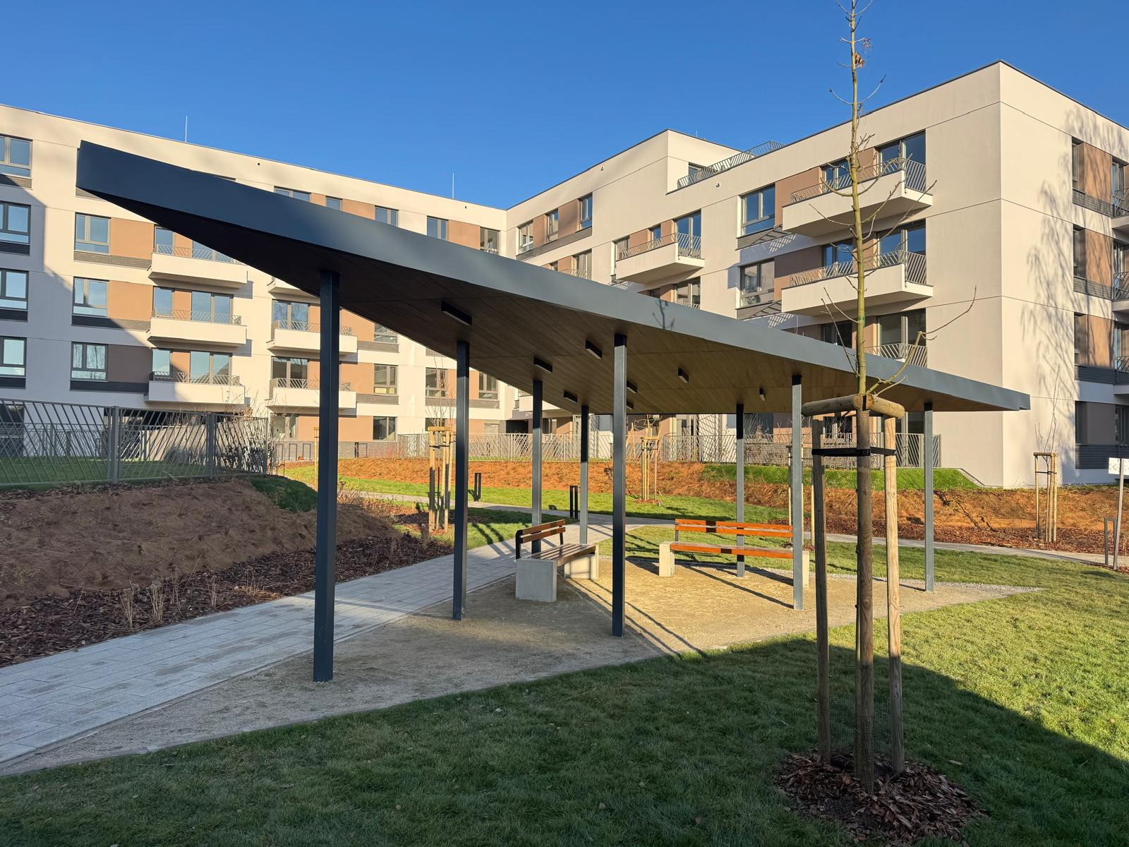 Nowoczesna, otwarta altana z szarą, stalową konstrukcją na terenie osiedla mieszkaniowego w słoneczny dzień. Widoczne ławki i trawnik.