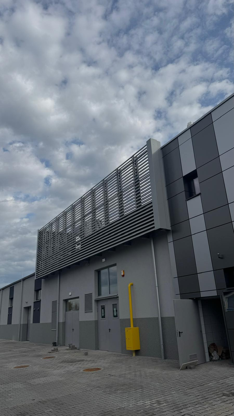 Nowoczesna hala z szarymi panelami i metalową konstrukcją na dachu, widok z dołu na tle pochmurnego nieba. Żółta instalacja gazowa przy ścianie.