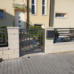 Nowoczesna, pozioma brama wjazdowa ze stali nierdzewnej, osadzona w niskim murku z jasnych bloczków, na tle domu z beżową elewacją i szarą kostką brukową.