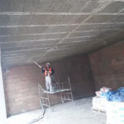 Pracownik w kasku i kamizelce na rusztowaniu nanosi zaprawę na sufit w pomieszczeniu z ceglanymi ścianami, widoczne materiały budowlane na palecie.