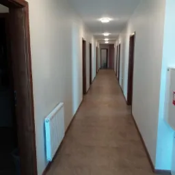 Długi korytarz z brązowymi drzwiami i beżowymi ścianami, oświetlony sufitowymi lampami, z grzejnikiem na ścianie po lewej stronie i brązową podłogą z płytek.