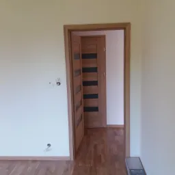 Wnętrze z drewnianą podłogą i dwiema drewnianymi ościeżnicami z drzwiami, jedne drzwi otwarte, drugie zamknięte, widoczne kable wystające ze ściany oraz paczka paneli podłogowych w rogu.