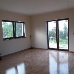 Wnętrze pokoju po malowaniu ścian na jasny kolor, z parkietem na podłodze, widoczne okno i drzwi balkonowe z widokiem na ogród, torba z narzędziami przy ścianie.